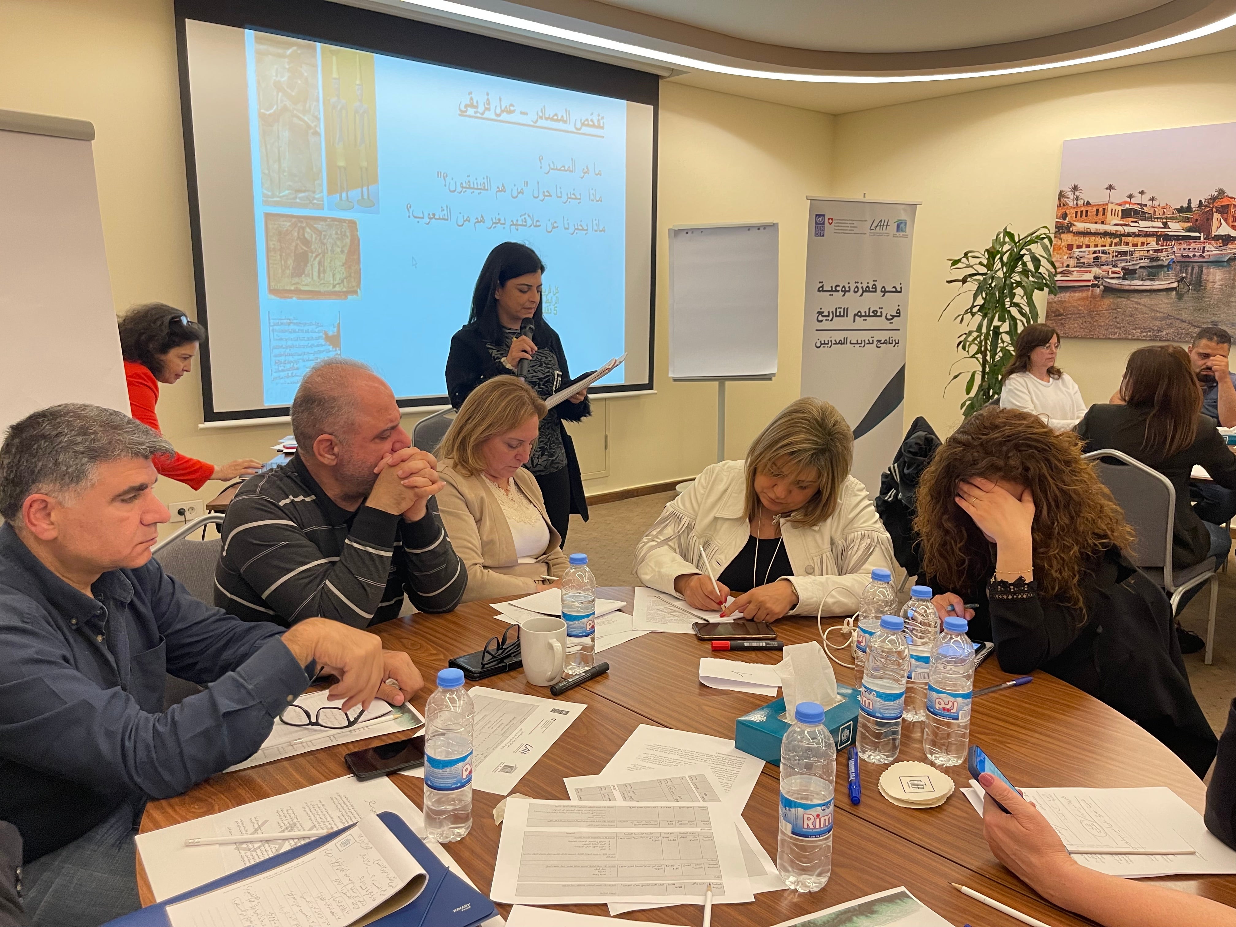 Participants gathered around table thinking and writing with trainer in the background