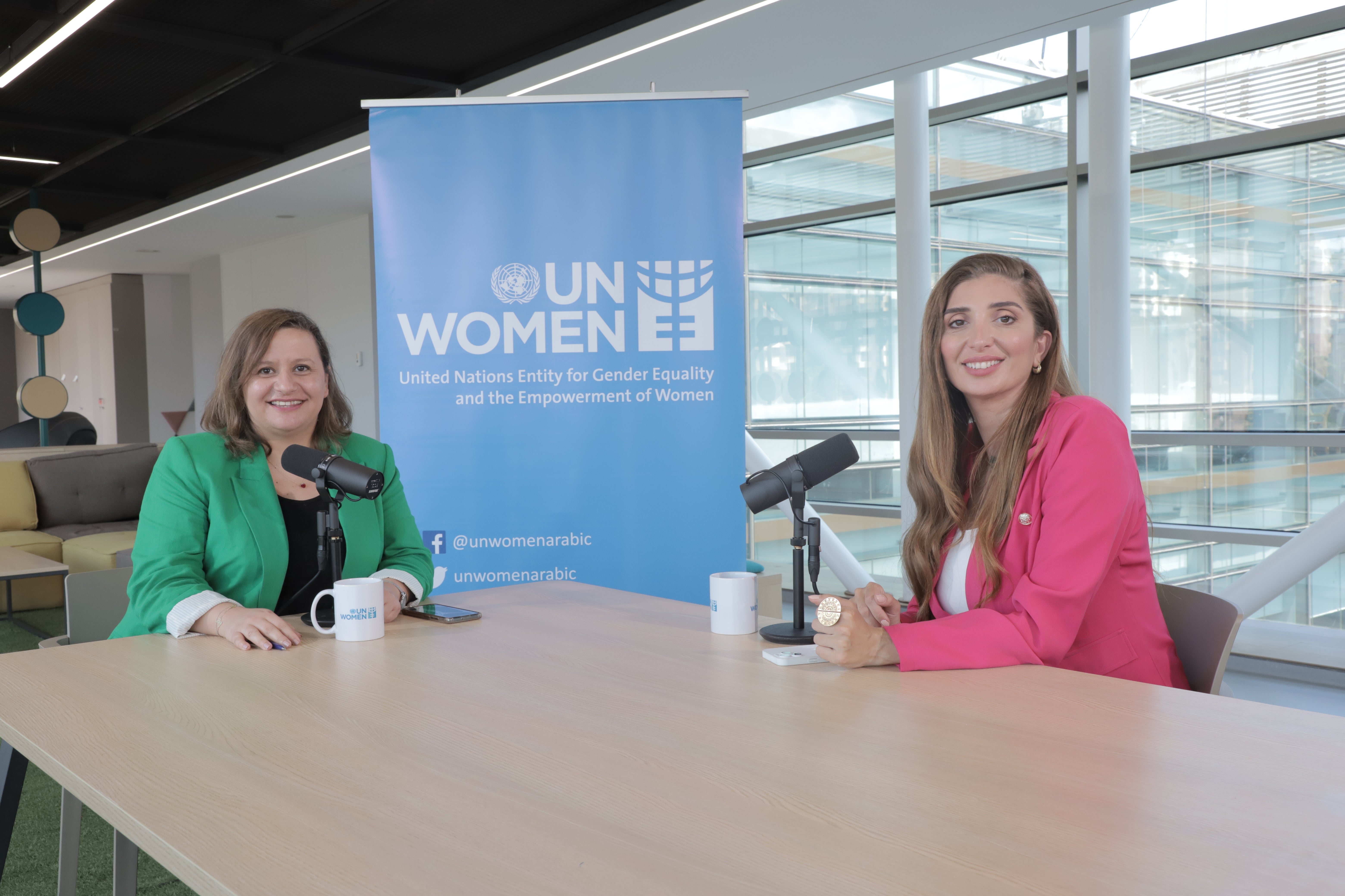 Two women facing the camera with microphones in front of them and UN Women logo in backgroung.