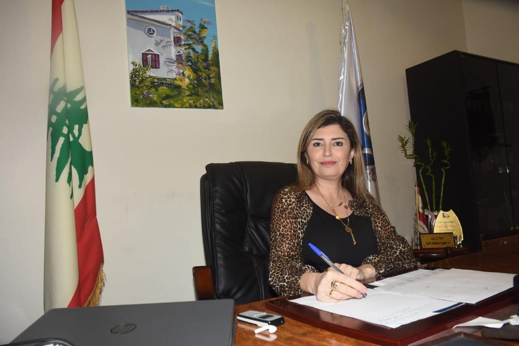 Woman sitting at desk with pen in hand and the Lebanese flag behind her.